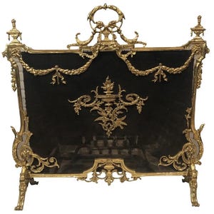 May include: A gold-toned metal fireplace screen with ornate detailing and a black mesh panel. The screen features a decorative design with floral motifs and a central urn.