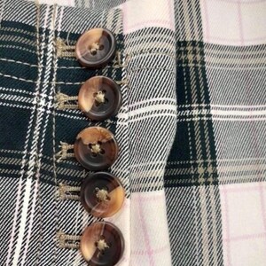 May include: Close-up of a plaid fabric with a buttoned closure. The fabric features a pattern of black, white, and green checks, with a pink accent. The buttons are brown with a two-tone effect.