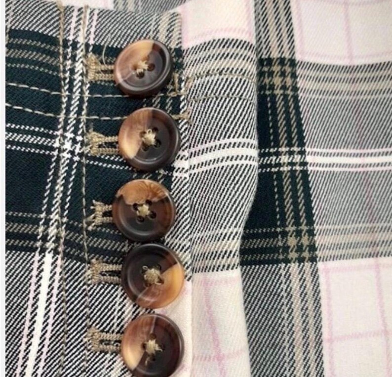 May include: Close-up of a plaid fabric with a buttoned closure. The fabric features a pattern of black, white, and green checks, with a pink accent. The buttons are brown with a two-tone effect.