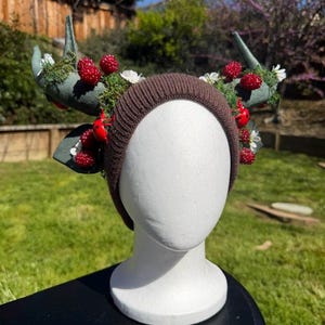 May include: A brown knit headband with decorative antlers, moss, red berries, and white flowers. The antlers are a muted green color. The headband is displayed on a white mannequin head.