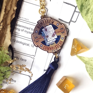 May include: A gold-colored keychain with a blue tassel and a round, colorful enamel pin featuring the words "Homebrew Connoisseur". The pin has a design of a book with a quill pen and stars.