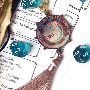 May include: A gold charm with a brown and white design that says "Backstory Club 1000+ 1K+" hanging from a gold chain with a star charm. The charm is surrounded by turquoise blue dice with white numbers.