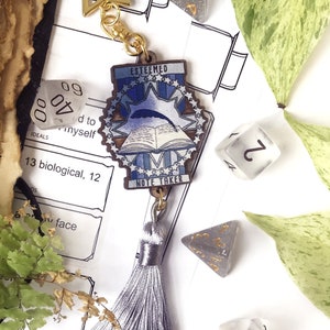 Könnte beinhalten: Ein blauer und weißer Holzanhänger mit den Worten "Esteemed Note Taker" und einem Buch mit einer Feder. Der Anhänger ist an einem silbernen Quasten und einer goldenen Kette mit einem Stern befestigt.