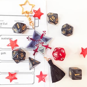 Puede incluir: Un surtido de dados de juego, amuletos rojos en forma de estrella y un llavero en forma de estrella con una borla negra. Los dados son negros con números dorados y un dado rojo con números blancos. Un amuleto dice "TPK VICTIM".
