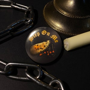 May include: A black button with a white and orange insect illustration and the text "Step On Me" in a white gothic font.