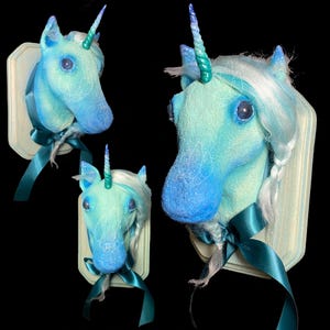 May include: Three blue and green unicorn head wall mounts with white manes and tails. Each unicorn head is mounted on a white wooden plaque with a teal ribbon.