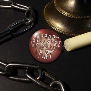 Puede incluir: Una chapa redonda con fondo rojo oscuro y texto blanco de estilo goteante que dice SUPPORT VAMPIRE (AND HUMAN) ART. La chapa está rodeada por una cadena de metal plateado y una vela de color crema con una base dorada.
