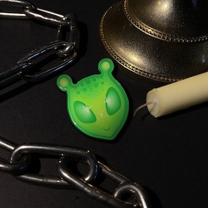May include: Green alien face pin with sparkly eyes and a smile. The pin is on a black background with a chain and a candle.