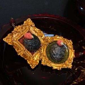 May include: Two ornate gold-colored framed pendants. Each frame surrounds a dark, textured stone with a small, pink accent. The frames have intricate detailing and are set against a dark red background. The pendants are connected by a chain.