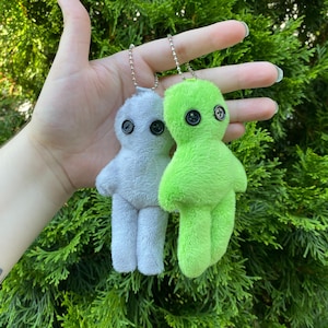 May include: Two plush dolls, one light gray and one bright green, each with button eyes and a chain. The dolls have a simple, rounded shape and are held against a backdrop of green foliage.
