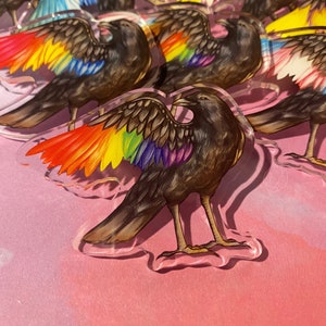 Puede incluir: Acr&iacute;lico transparente en forma de cuervo con alas de colores del arco&iacute;ris. Los cuervos tienen cuerpos marrones y est&aacute;n sobre una superficie rosa. Las alas muestran un espectro de colores vibrantes, incluyendo rojo, naranja, amarillo, verde, azul y morado.