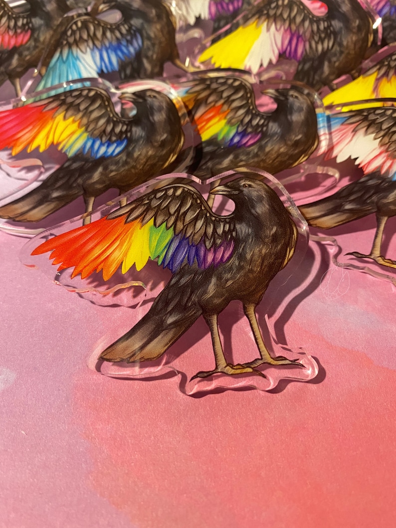 Puede incluir: Acr&iacute;lico transparente en forma de cuervo con alas de colores del arco&iacute;ris. Los cuervos tienen cuerpos marrones y est&aacute;n sobre una superficie rosa. Las alas muestran un espectro de colores vibrantes, incluyendo rojo, naranja, amarillo, verde, azul y morado.