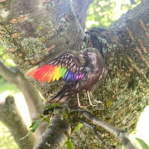 Puede incluir: Una pegatina transparente de un p&aacute;jaro con alas de arco&iacute;ris, posado en una rama de &aacute;rbol. El p&aacute;jaro tiene plumas marrones y grises y se encuentra sobre un fondo de corteza de &aacute;rbol y hojas verdes. La pegatina es transparente.