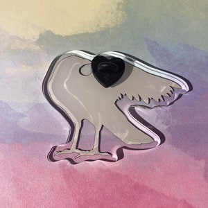 Puede incluir: Un pin de acr&iacute;lico transparente con forma de p&aacute;jaro con un respaldo de pin negro en forma de coraz&oacute;n. El p&aacute;jaro es de color gris claro con detalles grabados, sobre un fondo pastel de color rosa, morado y azul.