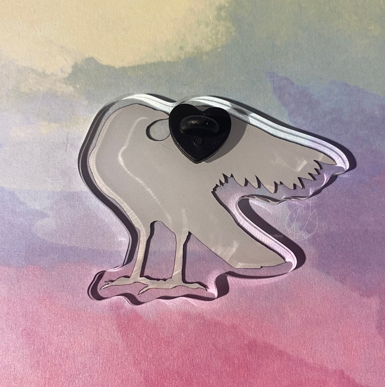Puede incluir: Un pin de acr&iacute;lico transparente con forma de p&aacute;jaro con un respaldo de pin negro en forma de coraz&oacute;n. El p&aacute;jaro es de color gris claro con detalles grabados, sobre un fondo pastel de color rosa, morado y azul.
