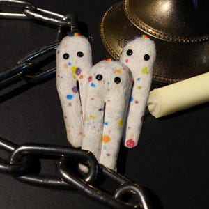 May include: Three white plush ghost figures with black button eyes and rainbow accents. The ghosts are covered in colorful paint splatters. A metal chain and a gold-colored bell are in the background.