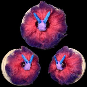 Può includere: Tre creature rotonde di peluche con un occhio centrale e antenne blu. Le creature hanno una forma soffice a forma di ciambella, con un gradiente di rosa e viola. La base è di colore beige chiaro e lo sfondo è nero.