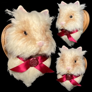 Puede incluir: Tres cabezas de conejo de peluche montadas sobre placas de madera. Los conejos tienen pelaje color crema con marcas bronceadas, narices rosas y ojos oscuros. Cada conejo lleva una cinta burdeos con un sello de cera. El fondo es negro.