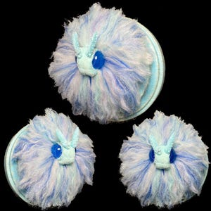 Op de afbeelding: Drie pluizige, ronde, lichtblauwe en witte decoratieve objecten die lijken op gestileerde insecten. Elk heeft een lichtblauw gezicht met grote blauwe ogen en antennes. De objecten zijn gemonteerd op lichtblauwe cirkelvormige bases.