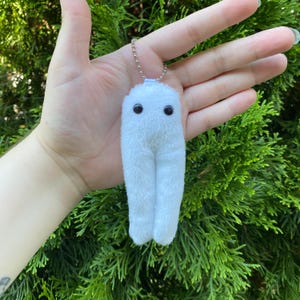 Könnte beinhalten: Ein weißer Plüsch-Schlüsselanhänger in Form eines kleinen, flauschigen Wesens mit zwei schwarzen Augen. Der Schlüsselanhänger ist an einer silbernen Kette befestigt. Der Hintergrund ist ein grüner Busch.