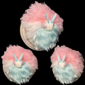 Può includere: Tre ornamenti soffici a forma di falena dai colori pastello. Ognuno ha una parte superiore rosa, un centro bianco e un fondo blu, con un viso bianco e antenne blu. Gli ornamenti sono su sfondo nero.