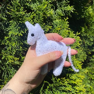 May include: A small, white, plush unicorn figurine featuring a textured surface with subtle, shimmering silver glitter accents. The unicorn has a single horn, a long, thin, white yarn tail, and a small, round, black bead eye on its side.