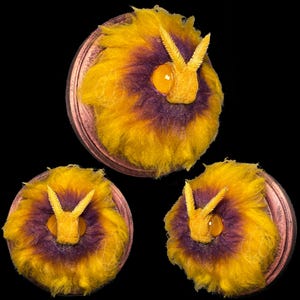 Puede incluir: Tres adornos de pared decorativos con un diseño central de pelaje amarillo y morado, que se asemeja a un insecto o criatura estilizada. Cada pieza tiene una base circular de color cobre y un elemento amarillo texturizado en el centro.