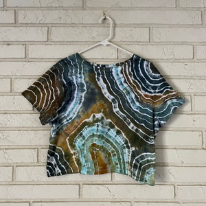 May include: A cropped, short-sleeved t-shirt with a tie-dye pattern in shades of brown, green, blue, and white. The pattern resembles a swirling, abstract design.