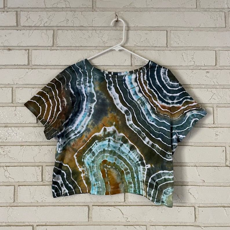 May include: A cropped, short-sleeved t-shirt with a tie-dye pattern in shades of brown, green, blue, and white. The pattern resembles a swirling, abstract design.