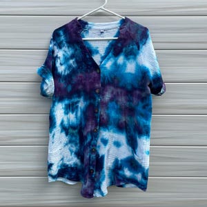 May include: A button-down shirt with a tie-dye pattern in shades of blue, purple, and white. The shirt has short sleeves, a V-neck, and two front pockets. It is hanging on a white hanger against a gray wooden background.