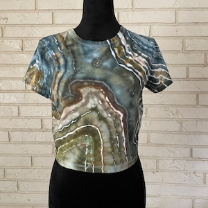 May include: A cropped, short-sleeved t-shirt with a tie-dye pattern in shades of green, brown, and blue. The pattern resembles agate or a similar stone.