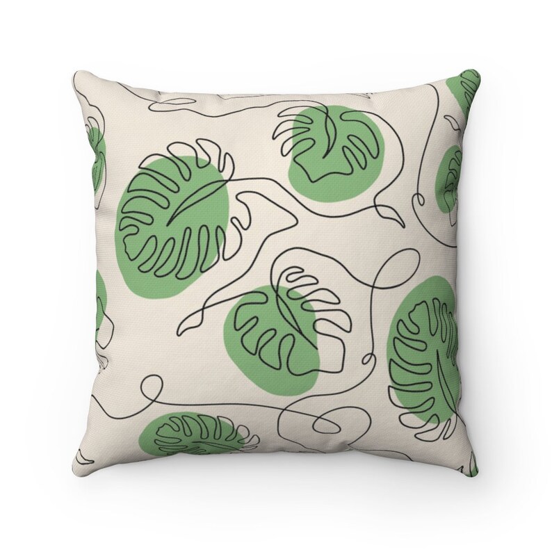May include: A throw pillow with a cream-colored background and a repeating pattern of green monstera leaves outlined in black. The leaves are stylized with a single line drawing.