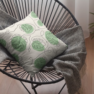 May include: A white throw pillow with a green leaf pattern. The pillow is on a black wire chair with a gray throw blanket.
