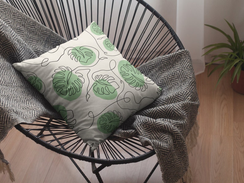 May include: A white throw pillow with a green leaf pattern. The pillow is on a black wire chair with a gray throw blanket.