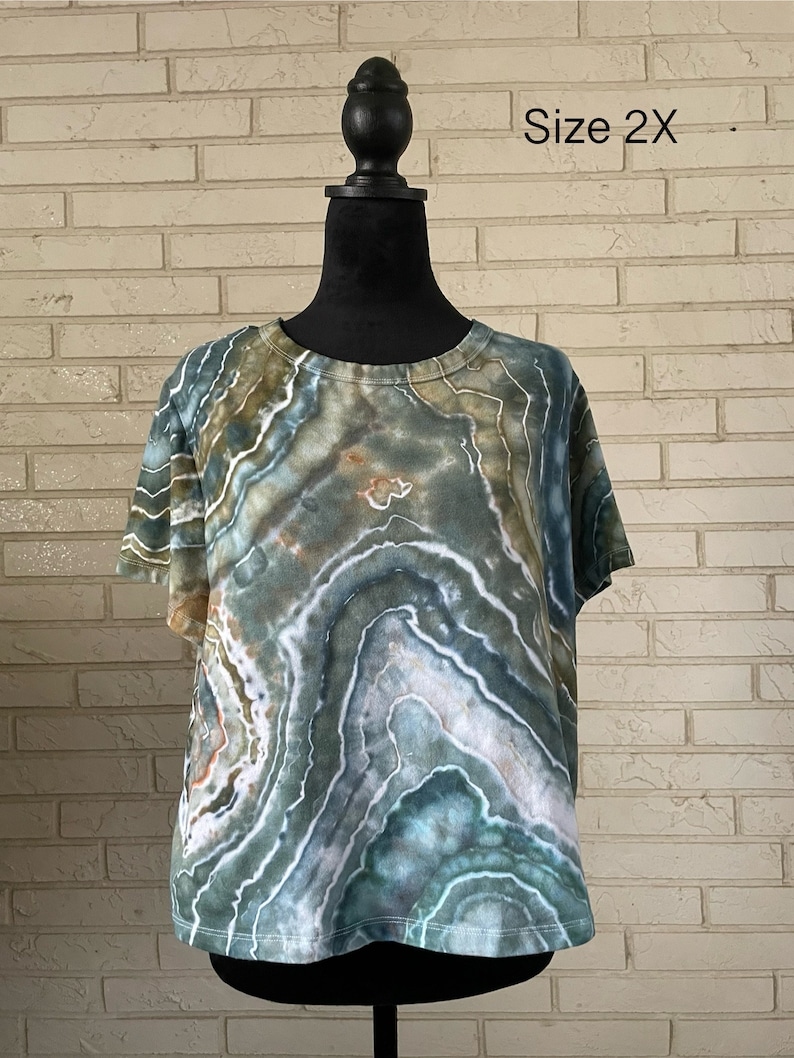 May include: A green, blue, and white tie-dye shirt with a round neckline and short sleeves. The shirt is on a mannequin and the text "Size 2X" is visible in the image.