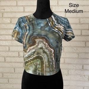 May include: A cropped, short-sleeved t-shirt with a tie-dye pattern in shades of green, brown, and white. The shirt has a round neckline and a relaxed fit. Size: Medium.