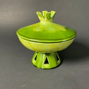May include: A ceramic, lidded serving dish in shades of green. The lid has a flower-like handle, and the base features triangular cutouts. The dish has a glossy finish and a vintage aesthetic.