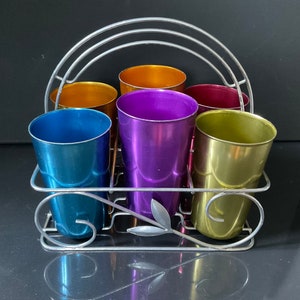 May include: A set of six colorful metal cups in a silver wire rack. The cups are in shades of blue, purple, orange, and yellow.