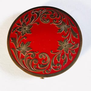 May include: A round, red compact with a gold-colored floral design. The compact has a gold-colored rim and a small clasp. The floral design includes flowers and leaves.
