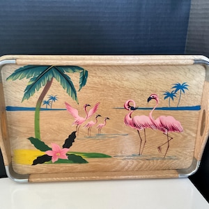 May include: Rectangular serving tray with a light wood grain pattern and metal trim. The tray features a painted tropical scene with pink flamingos, palm trees, and a pink flower. The tray has two handles.