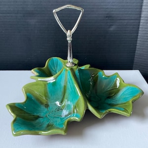 May include: A two-tiered serving dish with a leaf design. The dish is teal and green with a silver handle. The leaf-shaped bowls are connected at the center and have a glossy finish. The handle is a curved, open design.