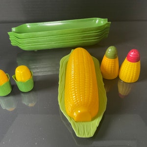 May include: A set of corn on the cob themed kitchenware, including a green serving dish, three yellow and red salt and pepper shakers, and two small yellow and green corn on the cob shaped containers.