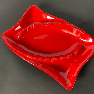 May include: A vibrant red ashtray with a unique, asymmetrical design. The ceramic ashtray features a glossy finish and several cigarette rests. This vintage piece is a stylish addition to any home decor.