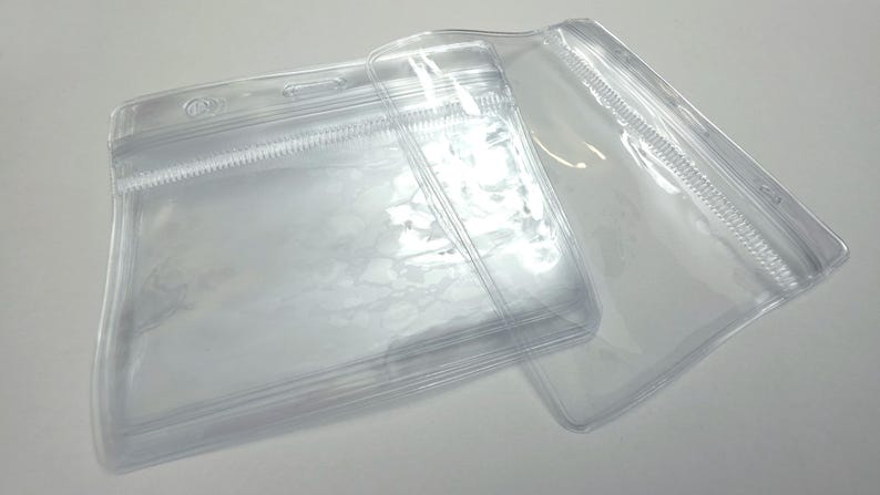 May include: Two clear plastic badge holders with horizontal orientation. Each holder features a reinforced top strip with a central slot for a lanyard or clip and a resealable zipper-style closure to protect the contents from dust and moisture.