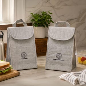 May include: Two gray insulated lunch bags with handles and flap closures. Each bag has a circular logo and text. One says "Pioneer Fuel" and the other says "Energía de Precursores". A sandwich and fruit are visible.