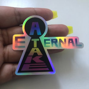 May include: Holographic sticker with the words "A ETERNAL TAKE" in a stylized design. The sticker has a black circle with the letter "A" at the top, and the rest of the text is in a gradient of colors. The sticker is held by a hand.