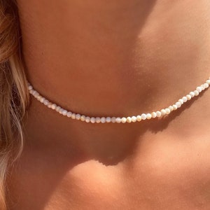 Può includere: Delicata collana girocollo composta da piccole perline rotonde bianche sporco. La collana è aderente al collo, creando un look minimalista ed elegante. Le perline hanno una lucentezza sottile, che riflette la luce. Un accessorio semplice ma elegante.