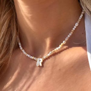 Puede incluir: Un collar de cuentas con pequeñas perlas irregulares de color blanco y crema intercaladas con cuentas diminutas. El collar incluye un colgante central prominente con la letra R, hecho de un material blanco liso e iridiscente.