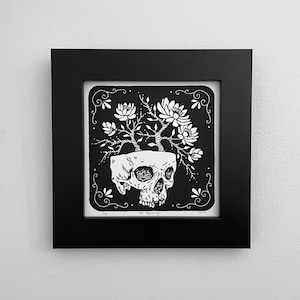 May include: Black and white print of a skull with flowers growing out of the top. The print is framed in black and has the text "New Beginnings" at the bottom.