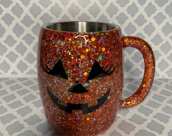 Può includere: Tazza di colore arancione con glitter e un design a forma di zucca nera. La tazza ha un interno in acciaio inossidabile e un manico.